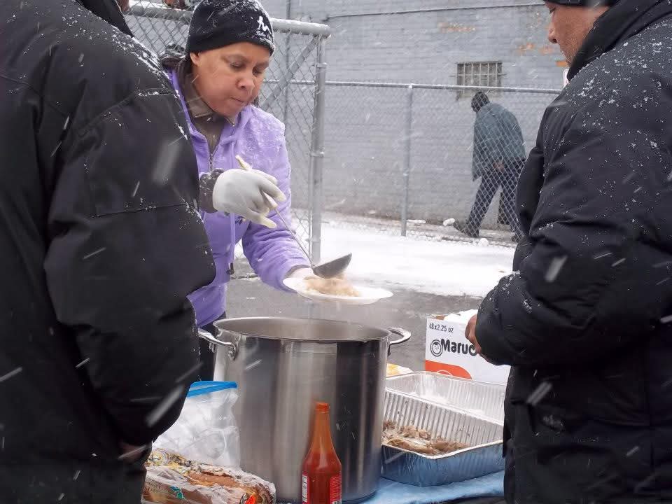 A volunteer in a lavender jacket ladles hot food from a stockpot onto plates during a snowy street outreach in Detroit