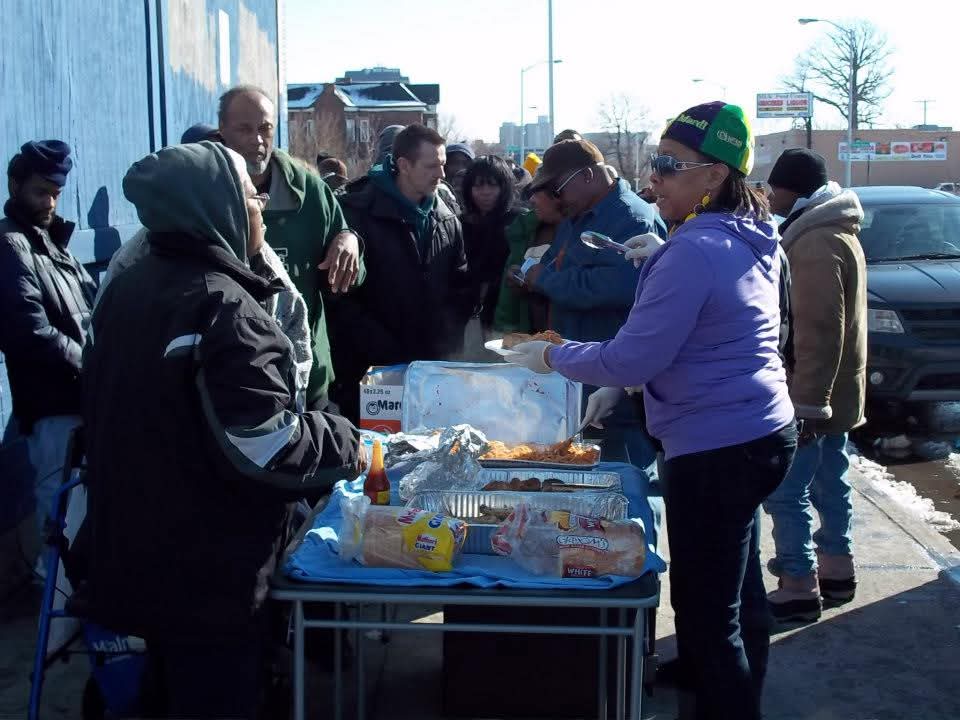 A woman in a purple jacket serves plates of pasta to a long line of neighbors beside a building on a winter day
