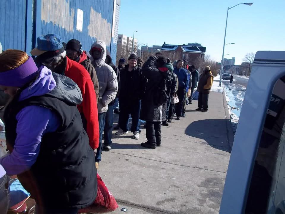 A long line of neighbors waits along a sidewalk beside a blue building on a winter day