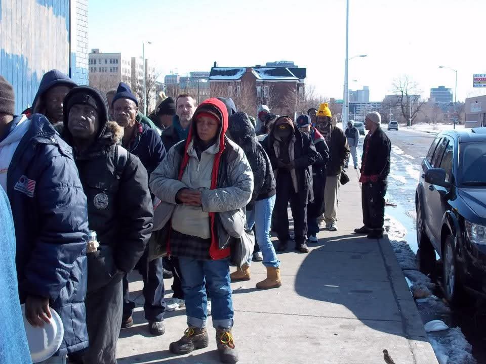 A line of people in winter coats waits along a Detroit sidewalk in the morning sun